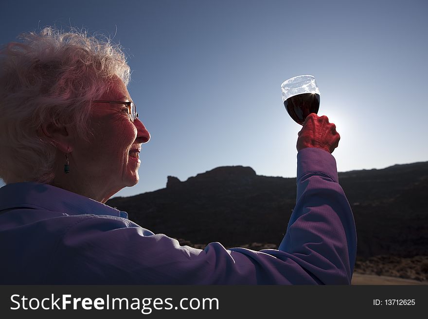 An elderly woman is in a desert landscape and holding up a glass of red wine in a toast. Horizontal shot. An elderly woman is in a desert landscape and holding up a glass of red wine in a toast. Horizontal shot.
