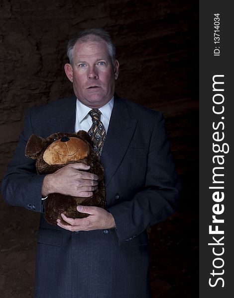 A businessman with an anxious expression is clutching a teddy bear for security. There is a rocky background seen behind. Vertical shot. A businessman with an anxious expression is clutching a teddy bear for security. There is a rocky background seen behind. Vertical shot.