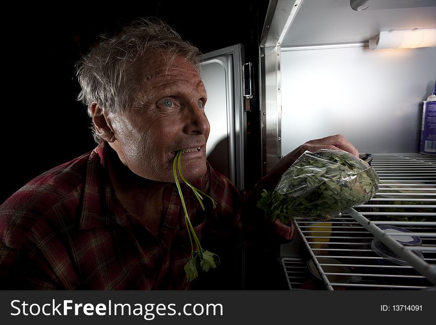 Side view of a crazed-looking middle-aged man staring intensely into a refrigerator that is mostly empty. He is holding a plastic bag of greens and also has some in his mouth. Horizontal format. Side view of a crazed-looking middle-aged man staring intensely into a refrigerator that is mostly empty. He is holding a plastic bag of greens and also has some in his mouth. Horizontal format.