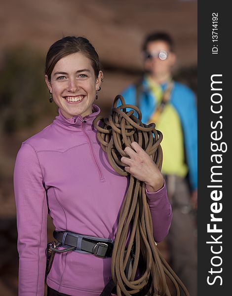 An attractive young woman is smiling and holding a climbing rope as her partner stands in the background. Vertical shot. An attractive young woman is smiling and holding a climbing rope as her partner stands in the background. Vertical shot.