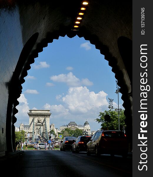 Perspective view of the Chain bridge in Budapest in bright sunlight from the Tunnel with car and passenger traffic and white clouds above.