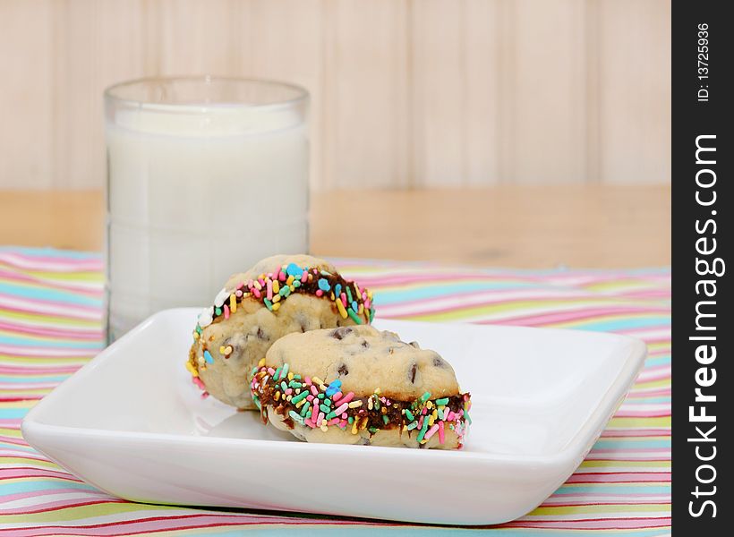 Plate with two chocolate chip cookie sandwiches with colorful sprinkles on a striped place mat with a glass of milk