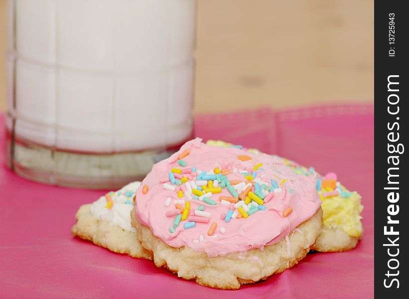 Three sugar cookies with icing and sprinkles with a glass of milk in the background on a pink place mat. Three sugar cookies with icing and sprinkles with a glass of milk in the background on a pink place mat