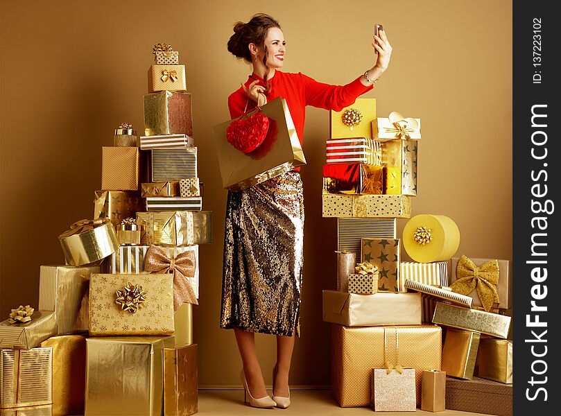 Full length portrait of happy trendy shopper woman in red blouse and golden sparkly sequin pencil skirt with shopping bag and red heart taking selfie with cellphone among 2 piles of golden gifts. Full length portrait of happy trendy shopper woman in red blouse and golden sparkly sequin pencil skirt with shopping bag and red heart taking selfie with cellphone among 2 piles of golden gifts