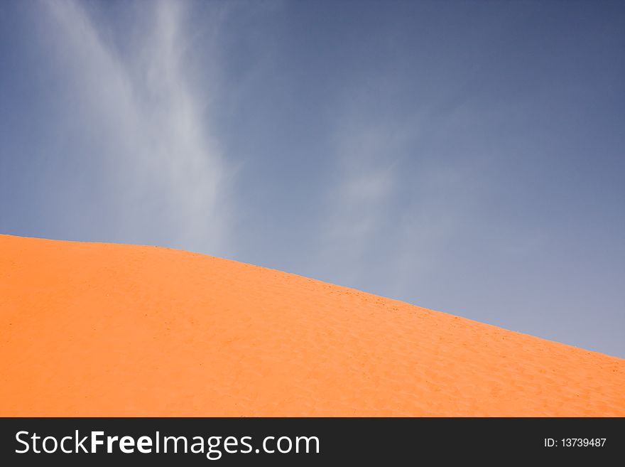 A featureless orange sand dune against a deep blue sky with whispy clouds