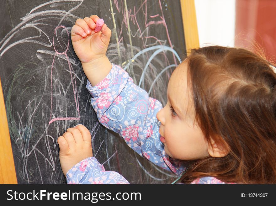 Cute little brown-haired girl is drawing on a blackboard. Cute little brown-haired girl is drawing on a blackboard