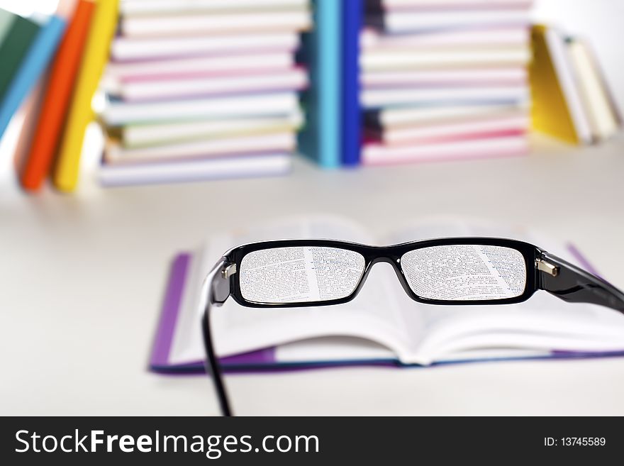 Clear view through specs facing open book in front of stack in rainbow colors paper wrapped books on white background, PHOTOGRAPH, NOT 3D RENDER.