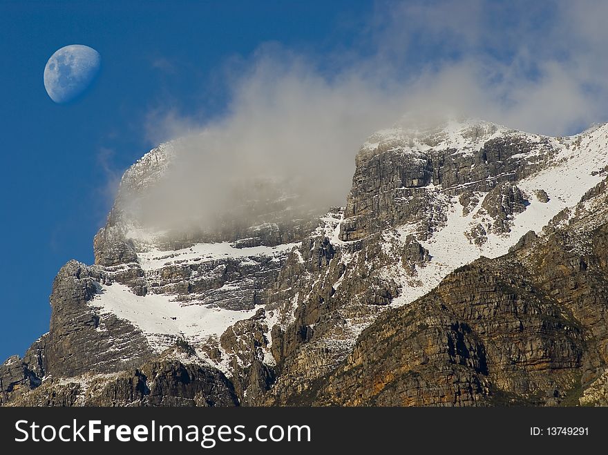 Snow covered mountain peak with clouds around it and the moon in the background