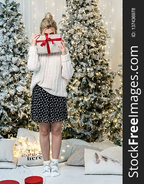 Blonde girl in white sweater hides the face behind the gift, christmas tree on background. New Year concept.