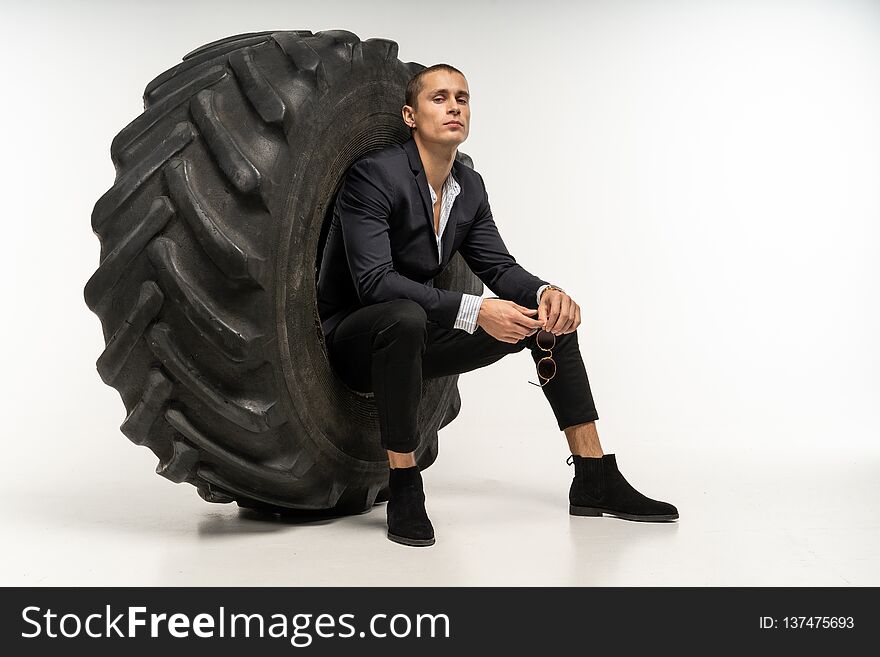 Stylish handsome man in black suit sitting in tire, posing in studio on white background