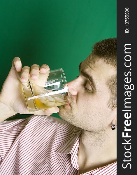 A young man dressed in a striped shirt on a green background makes the drink of whiskey on the rocks. A young man dressed in a striped shirt on a green background makes the drink of whiskey on the rocks