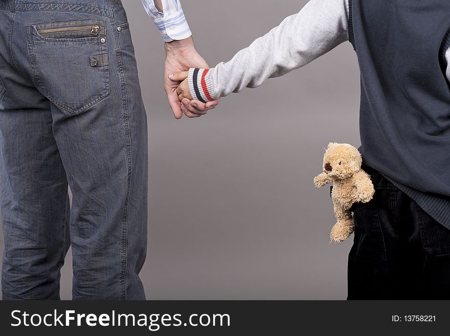 Young and careful father together with his teenager son holding hands of each other standing isolated over gray background with teddy bear. Young and careful father together with his teenager son holding hands of each other standing isolated over gray background with teddy bear