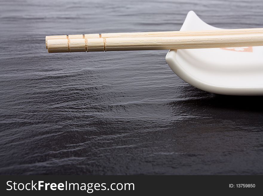 Pair of wood or bamboo Chopsticks in wrapper arranged on rest against dark slate background.