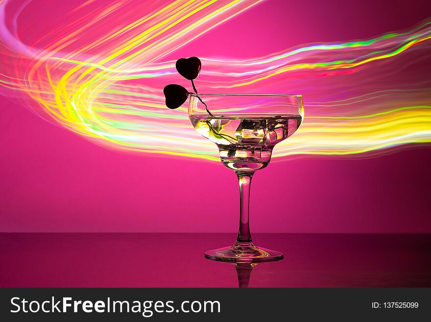 Glass with a cocktail and two hearts. Valentine`s day and wedding concept. On a dark pink background with light highlights