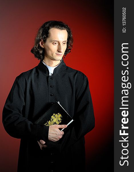 Conceptual portrait of Praying priest with wooden cross reading Holy Bible. red background