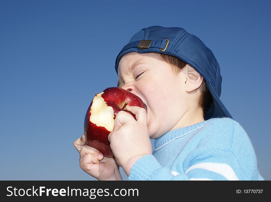 The child eagerly bites big red apple, blue cap, background sky. The child eagerly bites big red apple, blue cap, background sky