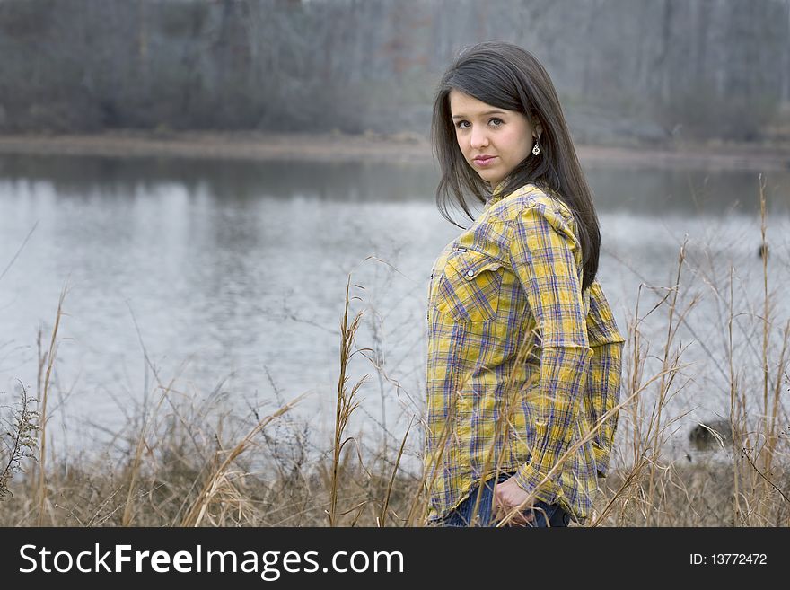 Beautiful young brunette woman with hands in pockets standing in front of lake. Beautiful young brunette woman with hands in pockets standing in front of lake