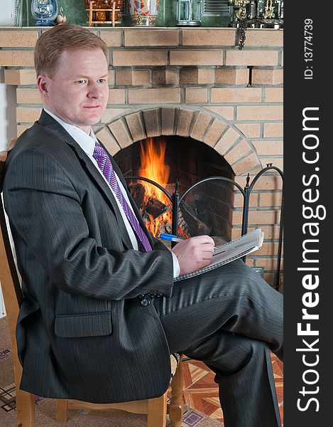 The man in a grey suit with a ball-point pen and a notebook at a house fireplace. The man in a grey suit with a ball-point pen and a notebook at a house fireplace.