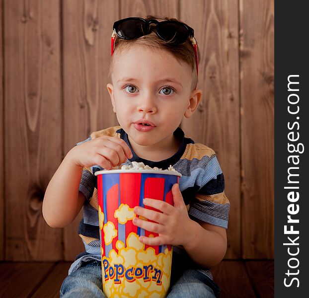 Little cute kid baby boy 2-3 years old , 3d cinema glasses holding bucket for popcorn, eating fast food on wooden background. Kids