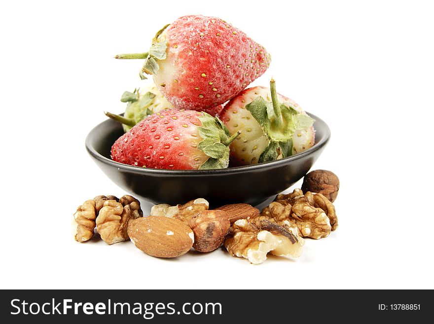 Red ripe frozen strawberries in a small black bowl with mixed nuts on a reflective white background. Red ripe frozen strawberries in a small black bowl with mixed nuts on a reflective white background
