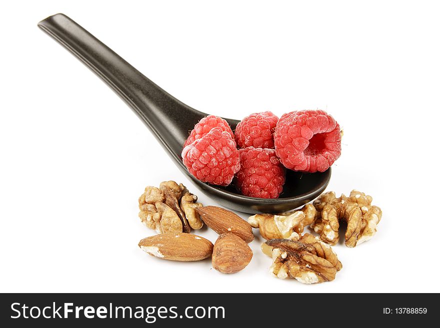 Red ripe frozen raspberries on a small black spoon with mixed nuts on a reflective white background. Red ripe frozen raspberries on a small black spoon with mixed nuts on a reflective white background