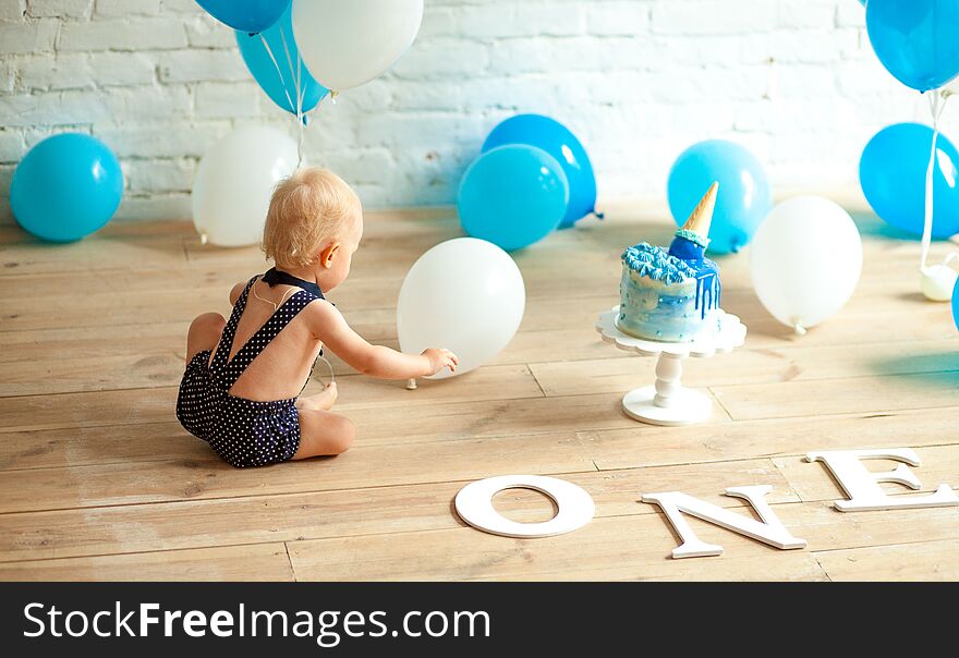 One year old boy is celebrating his first birthday among a lot of blue and white balloons, festive cake and inscription on floor. One year old boy is celebrating his first birthday among a lot of blue and white balloons, festive cake and inscription on floor