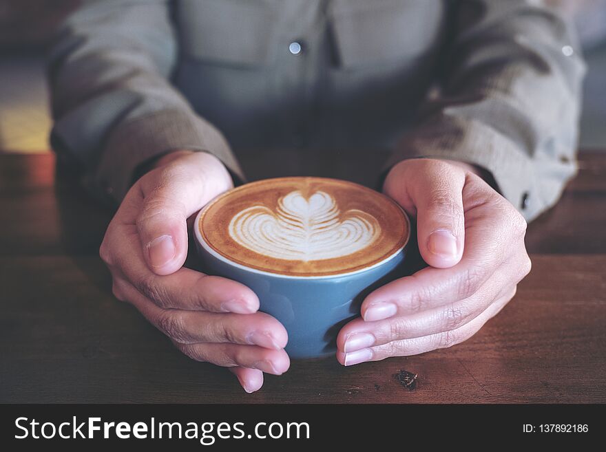 Closeup image of hands holding a blue cup of hot latte coffee with latte art on wooden table in cafe