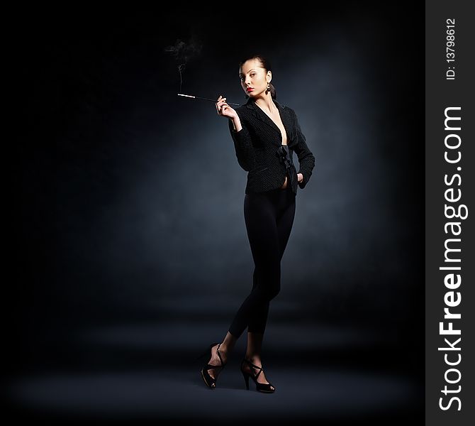 The portrait of a slim young lady in dark clothes holding a lighted cigarette in her hands. The portrait of a slim young lady in dark clothes holding a lighted cigarette in her hands