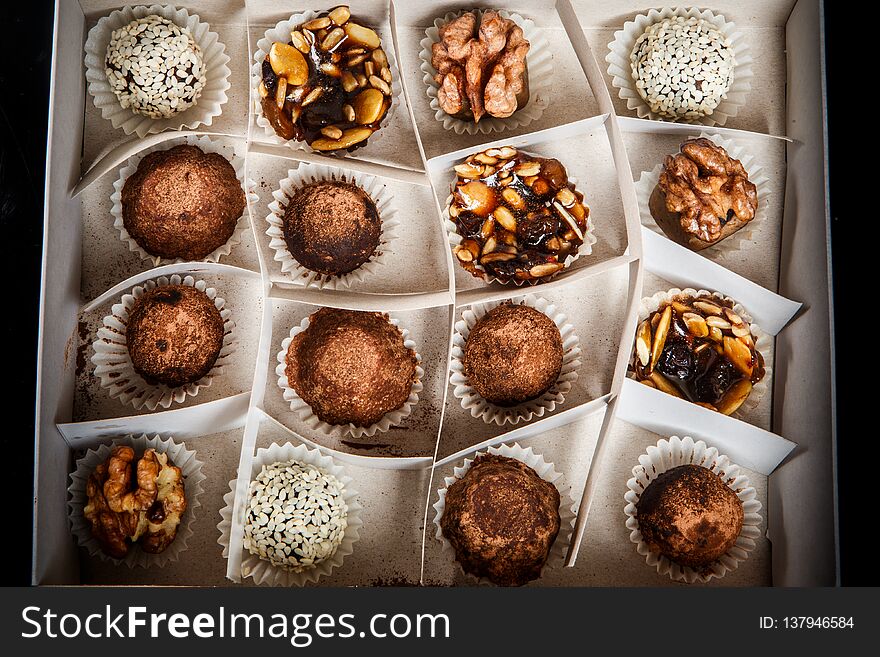 top view on big handmade box filled with assorted healthy useful handmade candies decorated with caramelized sunflower and pumpkin seeds, cocoa powder, walnut and sesame seeds on black mirror background