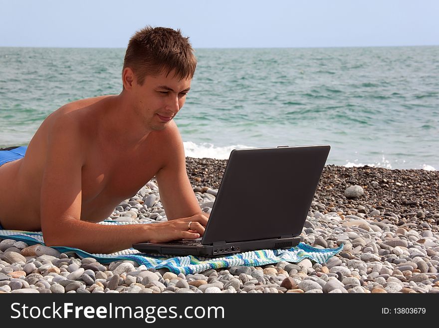Young man working on his notebook on seacoast. portrait of guy with laptop lying on beach.