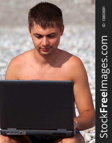 Young guy working on his notebook on seacoast. man with laptop sits on pebble beach.