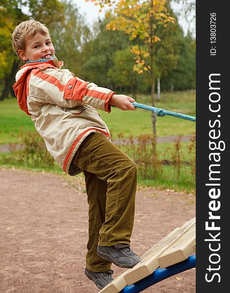 Boy in jacket is on playground in autumn park. Smiling, he climbs the stairs holding onto a rope.