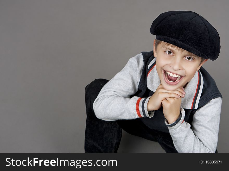 Young cute and smiling happy teenager boy sitting on floor with old cap and looking up having fun with mouth open and hands support chin isolated over gray background. Young cute and smiling happy teenager boy sitting on floor with old cap and looking up having fun with mouth open and hands support chin isolated over gray background