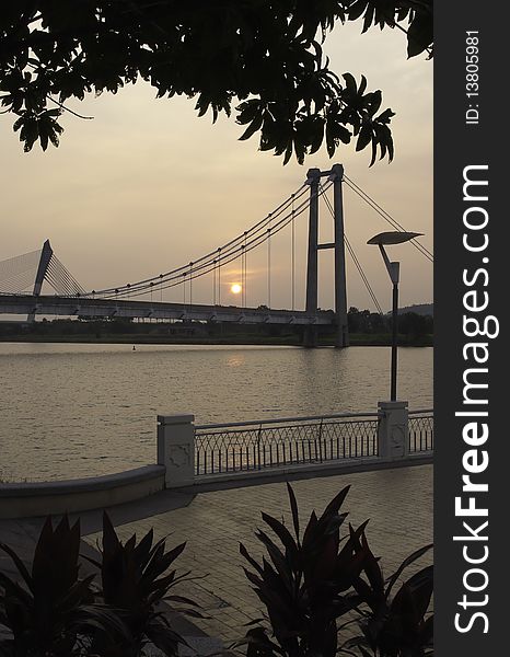 Scenic view of sunset seen in the middle of cable-stayed suspension bridge across lake framed by silhouette of tree and shrub on the lakefront. Scenic view of sunset seen in the middle of cable-stayed suspension bridge across lake framed by silhouette of tree and shrub on the lakefront