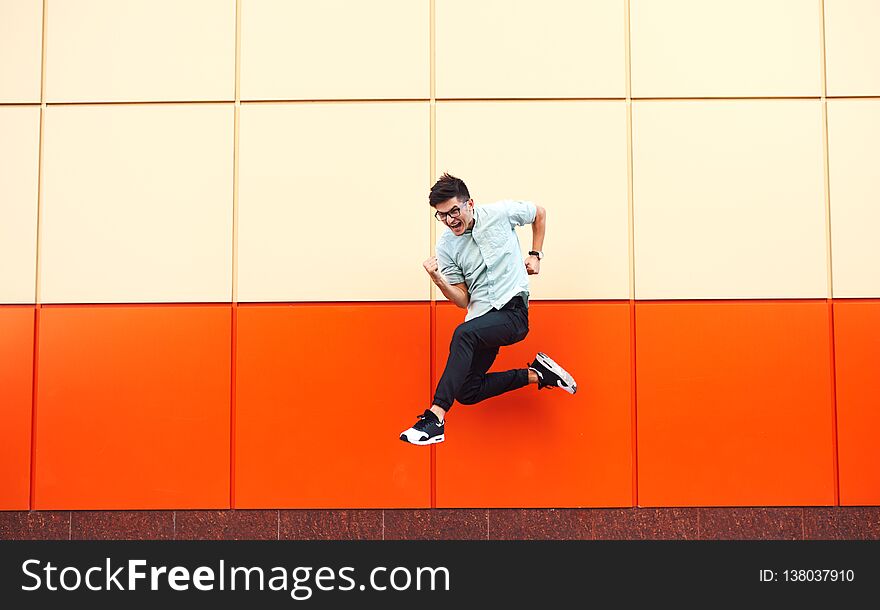 Pretty young man with glasses jumping against the colorful wall. Hipster in motion on ornage background