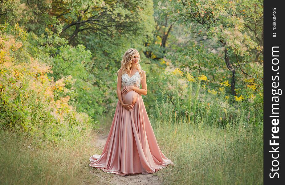 Young sweet pregnant girl with light curly hair hugs and holds her tummy, standing in a fairy forest in cold shades. dresses a long elegant satin silk pink dress with a lace top. art processing photo.