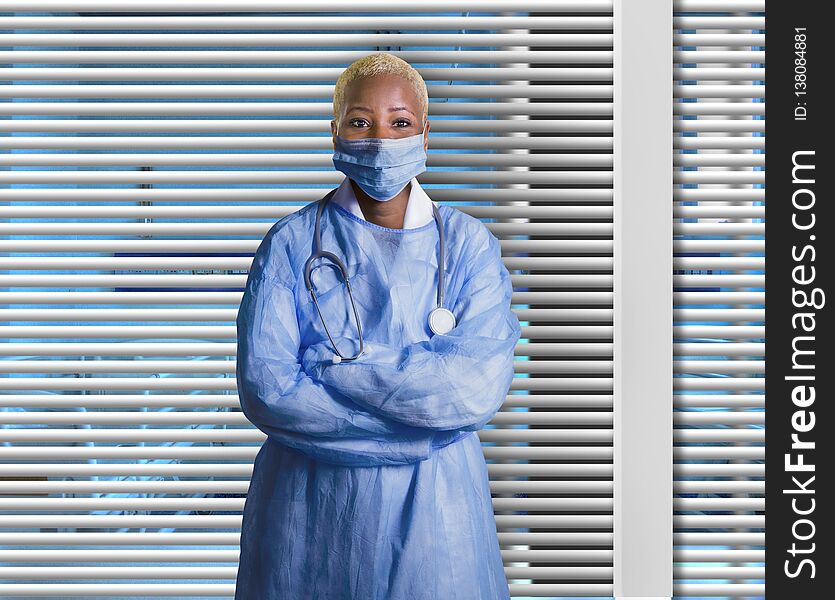 Young attractive and confident black African American medicine doctor wearing face mask and blue scrubs standing corporate at hospital room with Venetian blinds background