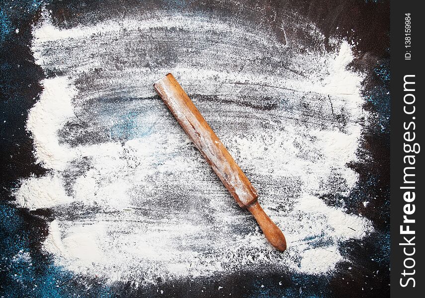 Food or baking background, white flour sprinkled on dark table, rustic wooden rolling pin, top view
