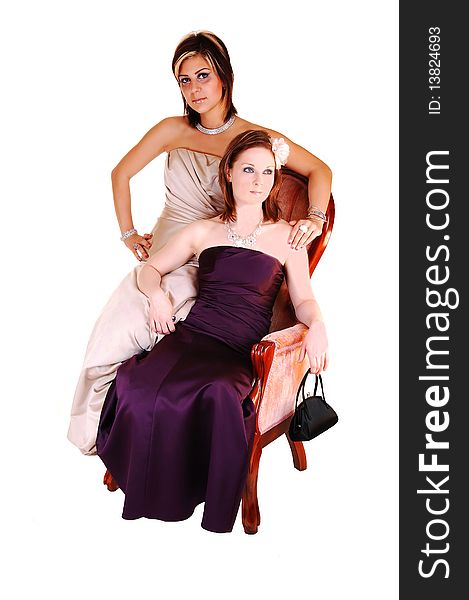 Two young girls and close friends, sitting in a pink antique armchair in long evening gowns, burgundy and beige, in the studio for white background. Two young girls and close friends, sitting in a pink antique armchair in long evening gowns, burgundy and beige, in the studio for white background.