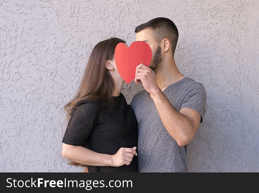 Waist-up portrait of sweet couple deeply in love sharing romantic kiss closing with red paper heart. Intimate moment. Side view. Isolated on grey background. Waist-up portrait of sweet couple deeply in love sharing romantic kiss closing with red paper heart. Intimate moment. Side view. Isolated on grey background