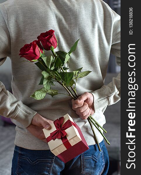 Young modern and elegant man holding red rose flowers and gift box behind his back at home. Surprised gift for woman. Romantic and valentines day holiday concept. Close up, selective focus. Young modern and elegant man holding red rose flowers and gift box behind his back at home. Surprised gift for woman. Romantic and valentines day holiday concept. Close up, selective focus