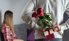 Mans Hands Hiding Holding Chic Bouquet Of Red Roses And Gift With White Ribbon Behind Back And Woman With Turned Head Awaits Surpr Stock Photo