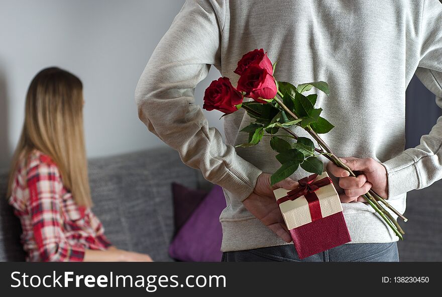 Mans hands hiding holding chic bouquet of red roses and gift with white ribbon behind back and woman with turned head awaits surpr