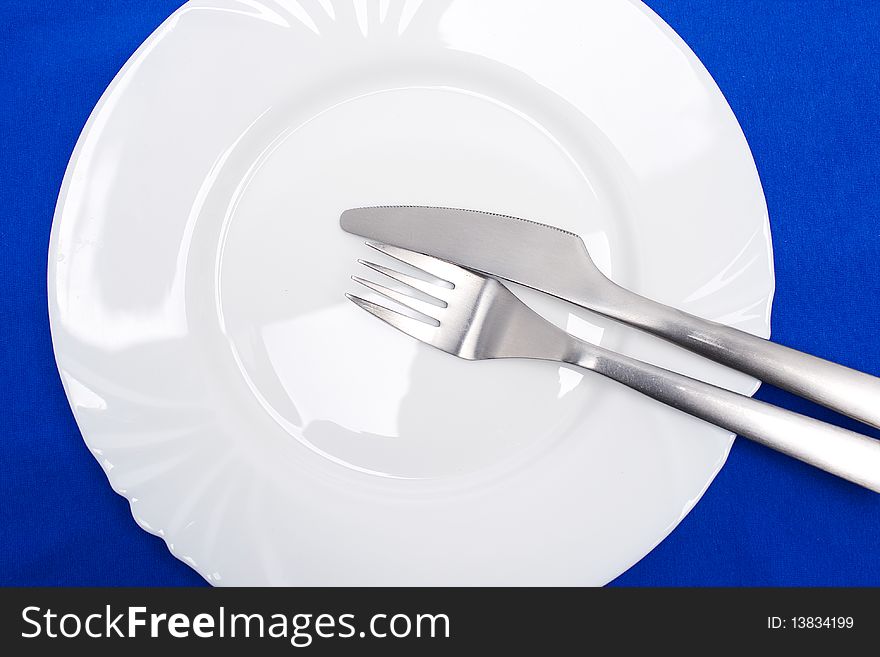 Fork and knife on top of a white plate over blue background. Fork and knife on top of a white plate over blue background