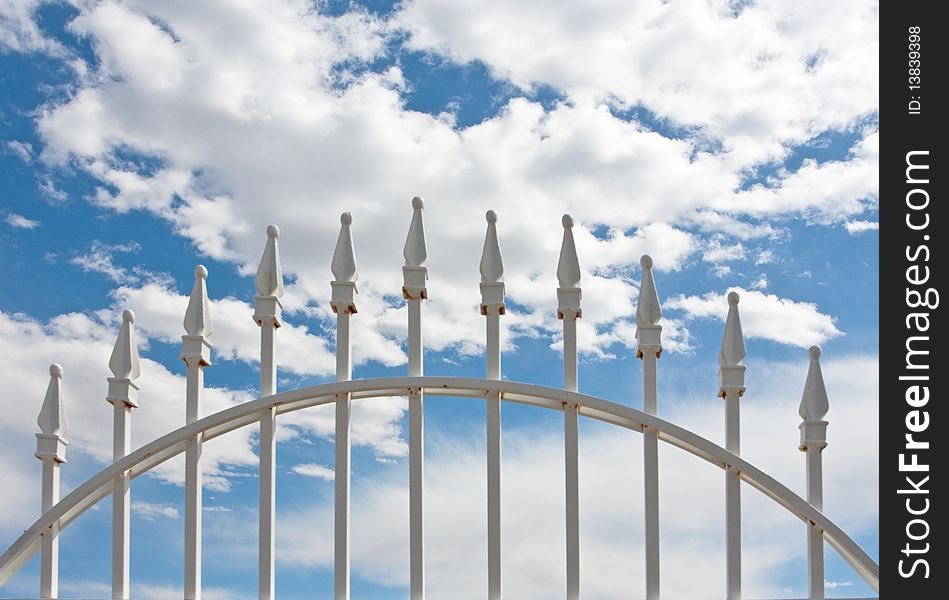 Detail of top of white wrought iron gate against blue sky and clouds