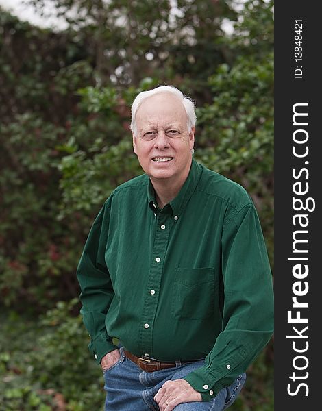Senior mature man wearing green shirt posing for photograph in front of green bushes.