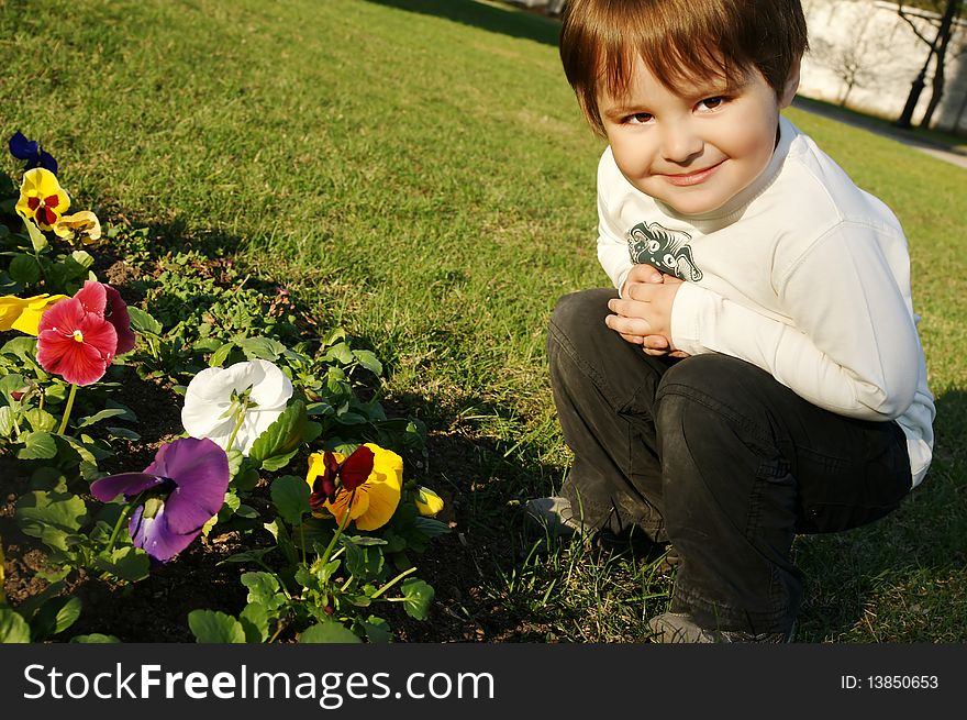 Beautiful smiling boy seating near flowers on green grass, outdoor shot