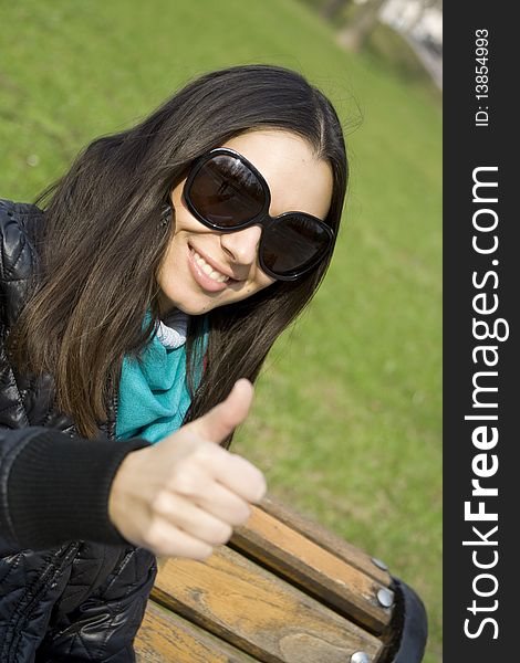 A beautiful young brunette girl in a park sitting on a bench smiling. Thumb sign OK. A beautiful young brunette girl in a park sitting on a bench smiling. Thumb sign OK