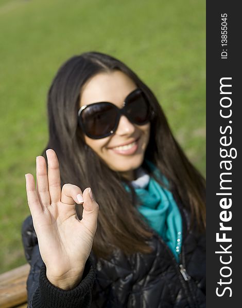 A beautiful young brunette girl in a park sitting on a bench smiling. Hand sign OK. A beautiful young brunette girl in a park sitting on a bench smiling. Hand sign OK