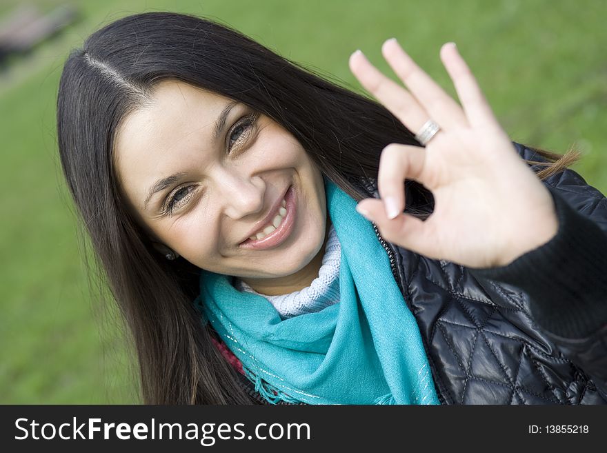 A beautiful young brunette girl in a park sitting on a bench smiling. Hand sign OK